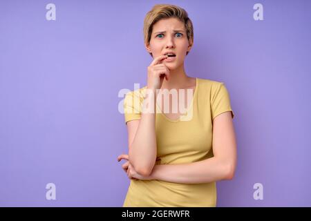 Confused short haired young woman touching mouth having dilemma difficult choice dressed in casual yellow t-shirt isolated over purple studio backgrou Stock Photo