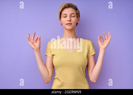 Young female in casual wear keep calm, meditating with eyes closed in studio on purple background, short-haired woman engaged in yoga, namaste. Nice l Stock Photo