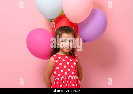 Gorgeous baby girl wearing summer pink polka dots dress, holding multicolored balloons behind her back, looking at camera, isolated over pink backgrou Stock Photo