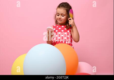 Beautiful Birthday girl in stylish pink dress with polka dots, combs her curly hair, admires her reflection on small pink cosmetic mirror. Portrait is Stock Photo