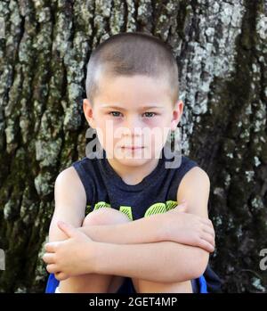 Little boy sitting outside against a tree, looks at camera unblinking.  His arms are crossed over his knees. Stock Photo