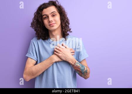 Kind caucasian male with long curly hair keep hands on chest feeling gratitude, thanks. man is looking at camera and smiling Stock Photo