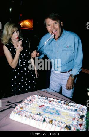 Glen Campbell pretends to lick the knife after cutting into his birthday cake at his surprise 60th birthday celebration on April 21, 1996 in Branson, Missouri. His wife, Kim looks on after sampling the icing on her fingers. Glen Campbell's actual birth date was April 22, 1936 Stock Photo