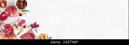Assortment of cheese and meat appetizers. Overhead view corner border on a white marble banner background with copy space. Stock Photo