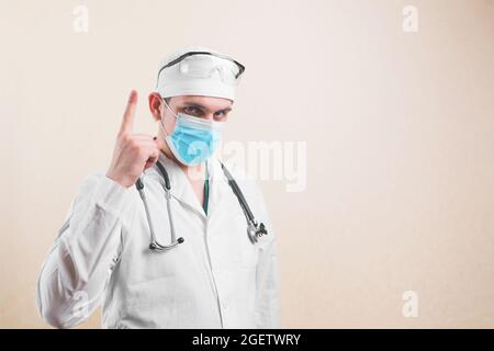 Young doctor in white lab coat, protective glasses and protective breathing mask pointing up on the yellow background. Stock Photo