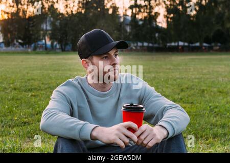 Young man drinking coffee from a plastic cup on the grass thoughtfully Stock Photo