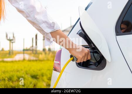 Female hands plugging in the charger into a socket of white electric car at a charging station near an electric power plant Stock Photo