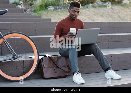 Young African student or freelancer with drink looking at laptop screen while sitting on staircase Stock Photo