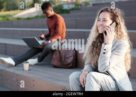 Blond businesswoman with smartphone by ear communicating against her male colleague outdoors Stock Photo