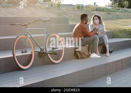 Two multuracial colleagues in masks having business discussion at meeting in urban environment Stock Photo