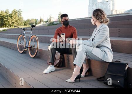Two young colleagues in protective masks enjoying break and interacting outdoors Stock Photo