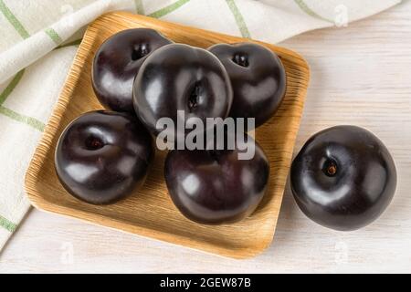 Ripe sweet black plums on a square wooden plate over white wooden table. Large fresh whole plums close-up. Ready to eat juicy fruits and berries. Stock Photo