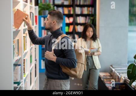 Adolescent man in casualwear taking book from bookshelf in college library Stock Photo