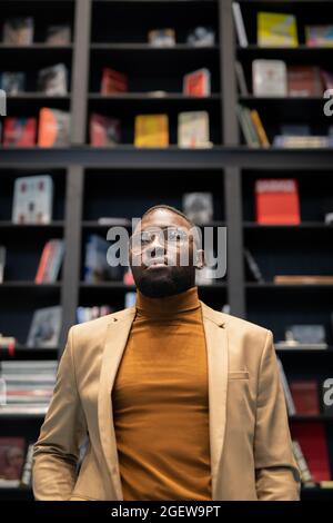 Young African-American man in smart casualwear standing in large bookshop against bookshelf Stock Photo