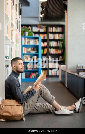 Young distracted man with open book looking aside while sitting on he floor against bookshelf Stock Photo