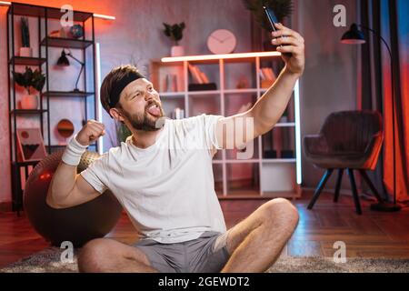 Leisure and recreation at home concept. Joyful handsome smiling young bearded guy in sportswear sitting on the floor with crossed legs, making selfie demonstrating his biceps and showing tongue Stock Photo