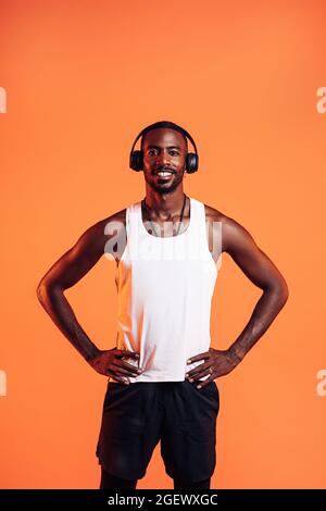 Happy guy wearing wireless headphones standing against an orange background with hands on his waist Stock Photo