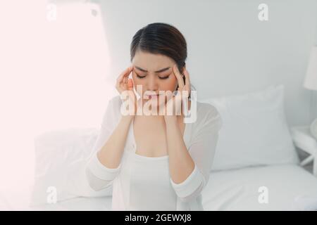 Miserable young woman touching temples sitting on bed at home, Stock Photo