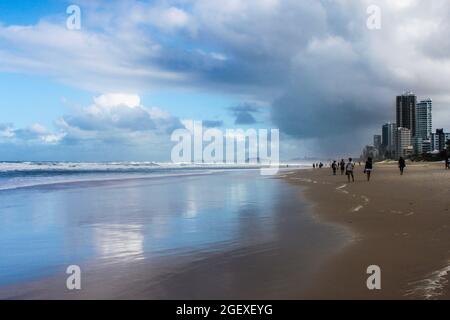 Tourists on beach under stormy sky and ocean reflecting the clouds and color and skyscrapers of the Gold Coast of Australia in the distance Stock Photo