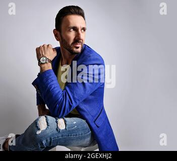 Portrait of mature unshaved man in modern torn jeans and blue jacket sitting on stool looking at copy space behind him  Stock Photo