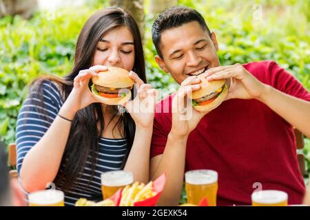 Happy couple eating hamburgers and drinking ice cold beers at an outdoor restaurant in a close up view of them smiling in anticipation as they bite in Stock Photo