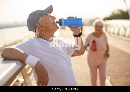 Positive senior man drinks water leaning onto railing while lady runs along footbridge Stock Photo