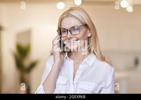 Pleasant talk. Attractive mature woman with blonde hair and glasses in casual wear talking on her phone, standing at home Stock Photo