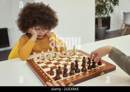 Cute player. Funny little boy with afro hair watching his friend making a move while playing chess, sitting at the table indoors Stock Photo