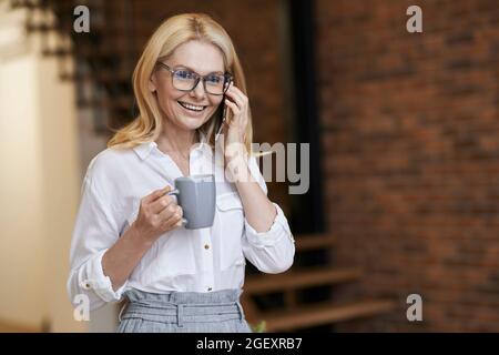 Nice day. Middle aged business woman with blonde hair and glasses holding a cup of coffee or tea while talking on her phone, standing at home Stock Photo
