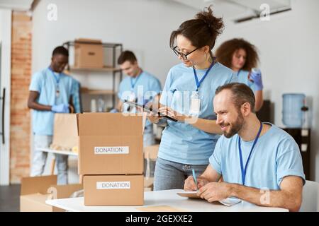 Man and woman happy to work together on donation project indoors. Team of volunteers in blue uniform making notes, calculating, sorting and packing Stock Photo