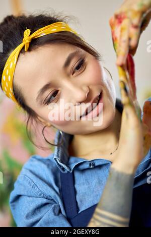Close up portrait of cheerful young woman, female painter looking at her hands in paint while creating painting in workshop Stock Photo