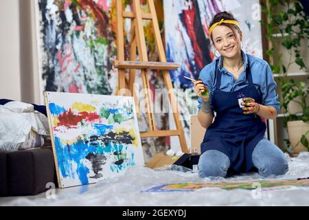 Joyful female painter in apron smiling at camera, holding paintbrush while working on painting, sitting on the floor at home studio workshop Stock Photo