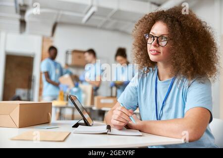 Young female volunteer in blue uniform looking away, making notes while working on donation project. Team sorting, packing items in the background Stock Photo