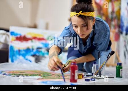 Focused young woman, female painter in apron choosing paint color, holding paintbrush while working on painting, sitting on the floor at home workshop Stock Photo