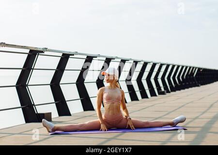 Side view of a young fitness woman, in sportswear, doing yoga exercises on the mat in the Front Splits pose while doing yoga on the seashore on the pi Stock Photo