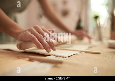 Crop anonymous female ceramist using slab roller to roll piece of clay on table with sticks and cutter while making handicraft pottery in studio Stock Photo