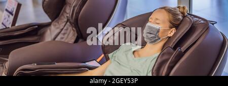 Beautiful young woman wearing a medical mask due to the coronavirus COVID 19 relaxing on the massage chair in airport or in the mall BANNER, LONG Stock Photo