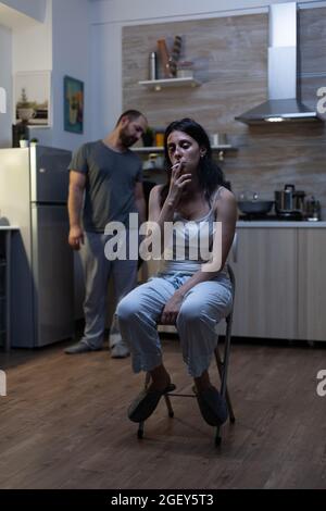 Unhappy terrified woman sitting and smoking cigarette feeling miserable after domestic violence and physical abuse from violent man. Aggressive drunk husband harassing bruised wife Stock Photo