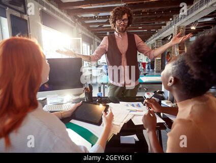 young caucasian beardy guy standing in front of the desk with his arms spread, smiling,  talking to his female colleagues sitting Stock Photo