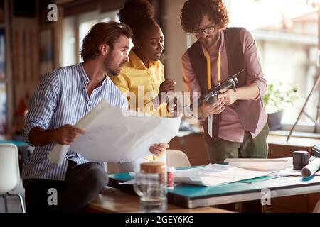 young beardy caucasian guy presenting drone as a solution for filming an advertisement to his male boss and afro-american female colleague Stock Photo