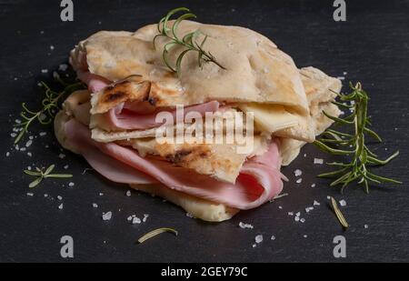 A piece of focaccia is garnished with slices of cheese and ham, decorated with sprigs of rosemary and coarse salt, on a slate base Stock Photo