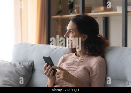 Smiling dreamy Latin woman sit on sofa holding modern cellphone Stock Photo