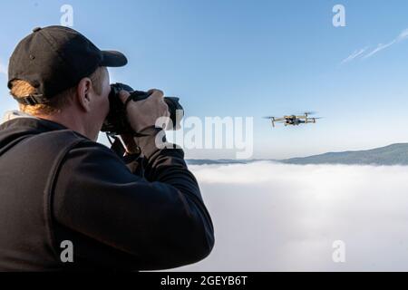 a male photographer takes pictures of a drone hovering in a thick fog in the air above the top of a mountain, while a copter takes it off. trips to na Stock Photo