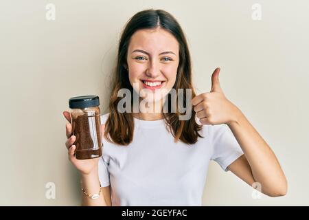 Young brunette woman holding soluble coffee smiling happy and positive, thumb up doing excellent and approval sign Stock Photo