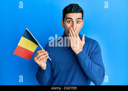 Handsome hispanic man holding belgium flag covering mouth with hand, shocked and afraid for mistake. surprised expression Stock Photo