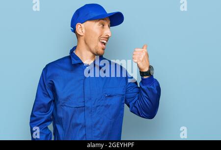Bald man with beard wearing builder jumpsuit uniform smiling with happy face looking and pointing to the side with thumb up. Stock Photo