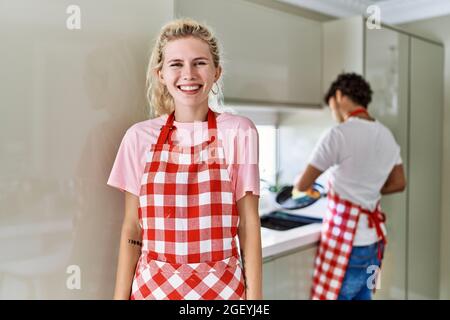 Young caucasian woman wearing apron and husband doing housework washing dishes looking positive and happy standing and smiling with a confident smile Stock Photo