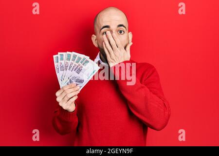 Young bald man holding egyptian pounds banknotes covering mouth with hand, shocked and afraid for mistake. surprised expression Stock Photo