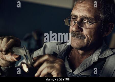An old man with glasses at work in the workshop measures a detail. Artistic photo of an elderly master. Cinematic processing of photography. Stock Photo