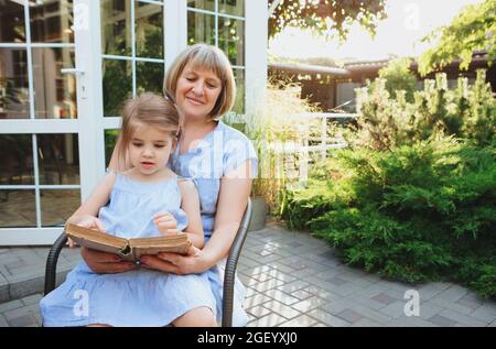 Senior grandmother and grandchild spending time together on sunny terrace near house, grandma reads fairy tales loud to lovely girl while little girl Stock Photo